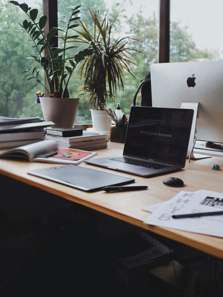 A laptop, books and notes on a desk beside a window.